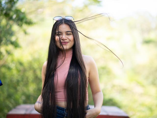 CamilaSofiia with long black hair, pink crop top, blue jeans, sunglasses on head, smiling, outdoors, green blurred background, wind blowing hair, hands on hips, natural light.