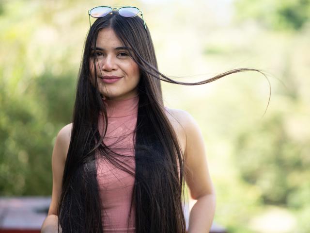 CamilaSofiia has long black hair, a pink sleeveless top, and a slight smile, with sunglasses on her head, and her hair flying to the right.