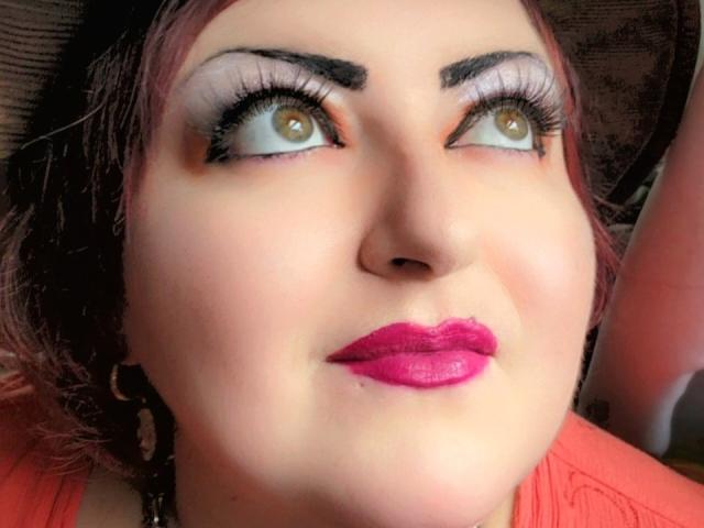 SkepticalIluzions has pink lipstick, dark red eyeshadow, heavy black eyeliner, and thick eyebrows. She's looking up with brown eyes, and has red-dyed hair. Her face is centered.