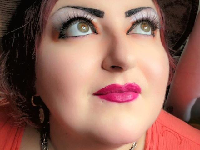 SkepticalIluzions a la peau pâle, du rouge à lèvres rouge, des sourcils noirs épais et des yeux jaunes qui regardent vers le haut. Elle porte un haut rose, a les cheveux foncés et porte des boucles d'oreilles noires..