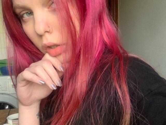 Lialit, con i capelli rosa-rossi, un anello al naso e la pelle pallida, morde il dito con le unghie viola, indossa una maglietta nera e sembra leggermente infastidita. I suoi seni sono parzialmente visibili..