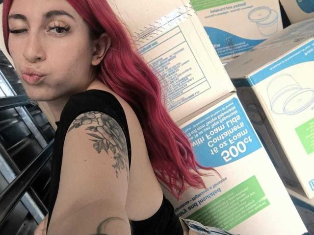 SammyViolet with red hair, winking, black top, tattoo on right arm, pouty lips, looking at camera, surrounded by stacked boxes, visible from side, showing back and profile.