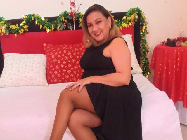 MatureYayita with blonde hair, wearing a black dress, sitting on a bed with white sheets, exposing her thighs, and smiling, with Christmas decorations in the background.