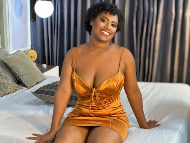 NinaCooper with curly black hair, brown skin, and a cute smile, sits on a bed in an orange, low-cut, velvet dress, showing her big, round, shiny boobs.
