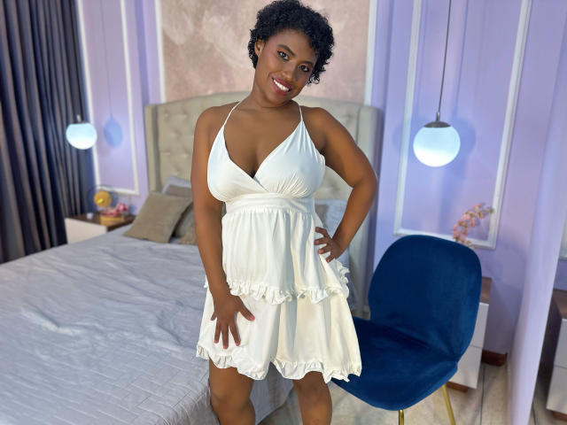 NinaCooper with dark skin, curly black hair, and a white halter dress showing her boobs and ass, smiling, hands on hips, standing in a bedroom.