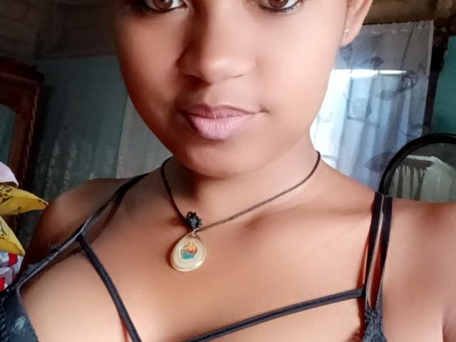 MeriAndMark's face, brown skin, small nose, pink lips, wearing a black strappy top, necklace with a pendant, subtle makeup, and a slight smile. Boobs visible, cleavage.