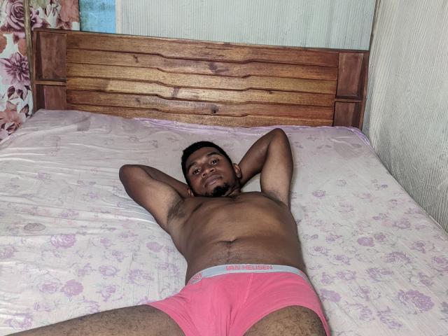 MeriAndMark is lying on a bed, shirtless, wearing pink boxer shorts, arms behind head, looking at camera, with a dark skin tone and short beard.