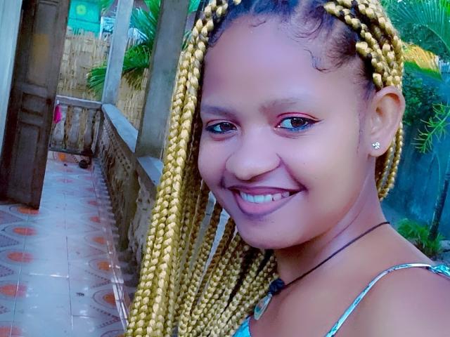 MeriAndMark has braided yellow hair, a dark brown skin tone, and a cute smile with visible teeth. She's wearing a necklace and has small earrings. Her tits are not visible.