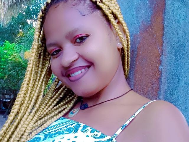 MeriAndMark with blonde braids, smiling with pink eyeshadow, showing white teeth, wearing a patterned top, necklace with a blue stone, against a textured wall.