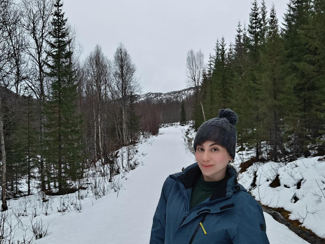 LuciaPerth has pale skin, dark eyebrows, and is wearing a blue jacket, gray beanie, and green shirt, standing on a snowy forest path with green and brown trees.