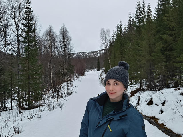 LuciaPerth with a cute face, green eyes, and a slight smile, wearing a gray knit beanie and blue jacket, standing in a snowy forest with pine trees.