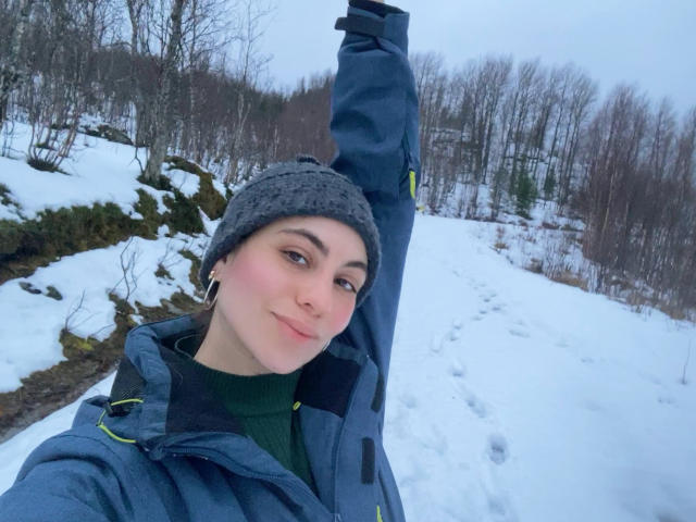LuciaPerth with raised arm, wearing a gray beanie, green turtleneck, and blue jacket, smiling in a snowy landscape. She has pink cheeks and hoop earrings.