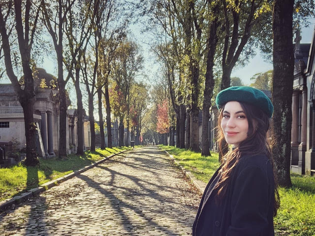 LuciaPerth with green beret, dark wavy hair, and black jacket, standing on sunlit cobblestone path, surrounded by trees, looking back with a slight smile.
