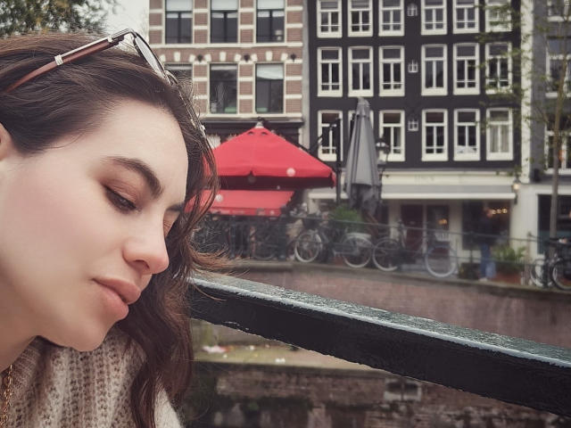LuciaPerth with brown wavy hair, light makeup, and a pink sweater, eyes closed, resting on a green railing, sunglasses on head, booru-style, wet railing, red umbrellas in background.