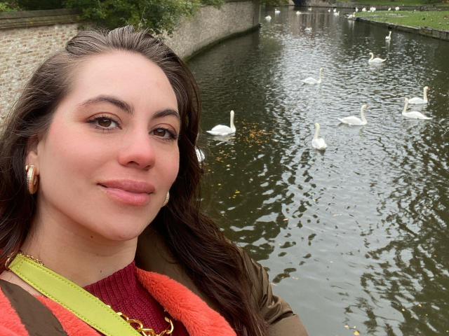 LuciaPerth has a cute face with brown eyes, wearing large gold earrings and a green-yellow strap, standing by a pond with white swans. Her brown hair is down.
