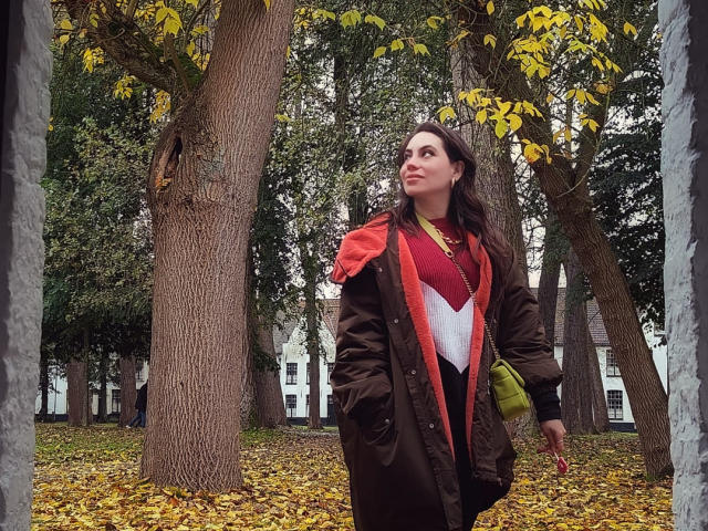 LuciaPerth with medium tits, wearing a brown coat with red and white pattern, green bag, and yellow leaves in background, looking up, standing in front of tall trees.