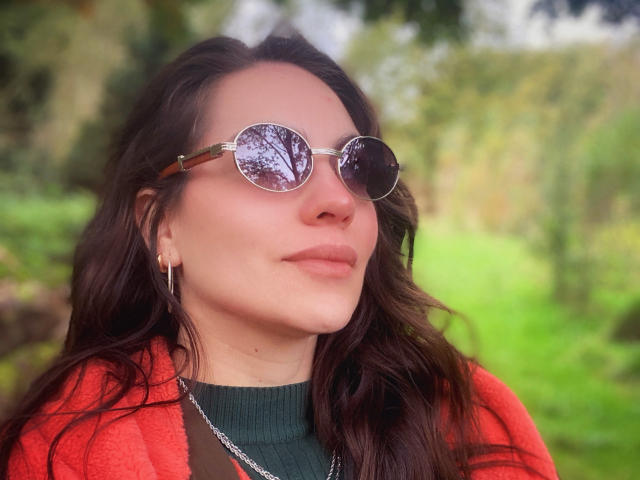 LuciaPerth with round, reflective sunglasses, dark wavy hair, red cardigan, green turtleneck, silver hoop earrings, and a slight smile, standing outdoors with blurred greenery in the background.