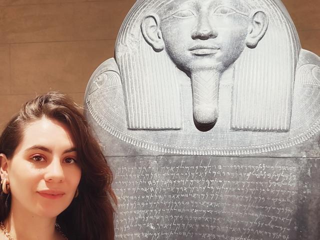 LuciaPerth with brown wavy hair, wearing earrings, stands next to a large stone bust of an Egyptian-looking face with a calm expression. Boobs not visible. Pussy not visible.