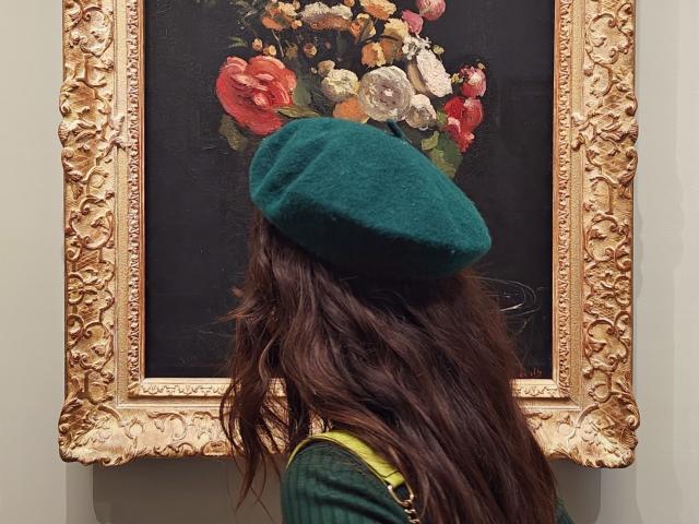 LuciaPerth with wavy brown hair, green beret, green shirt, and yellow strap, standing in front of an ornate, floral painting. Boob-sized beret, flowers in painting, gold frame.