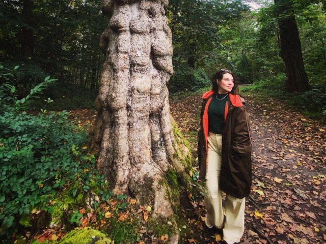 LuciaPerth stands next to a massive, textured tree with a rough bole, wearing a brown coat with a red collar, green shirt, and beige pants in a forest.