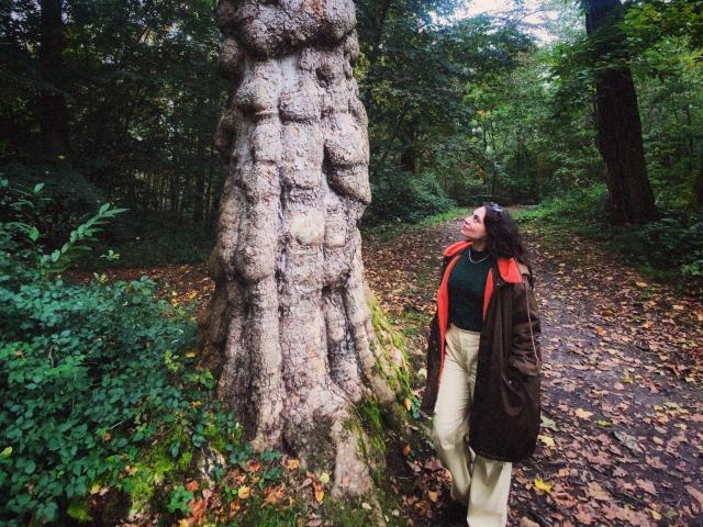 LuciaPerth stands next to a huge tree with gnarled, textured trunk. She's wearing a brown jacket, red scarf, and beige pants, looking up with a slight smile.