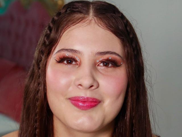 KathyStone has brown braided hair, pink lipstick, and eye makeup with small sparkling jewels above her eyebrows. She's smiling with pink glossy lips and a fair skin tone.
