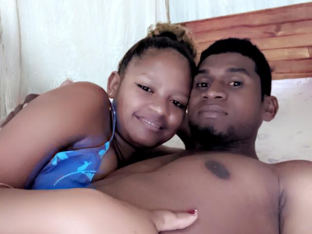 MeriAndMark with dark skin, curly hair, and small tits, smiling, lying on bed, next to shirtless MeriAndMark with black beard and small nipple, blue patterned top.