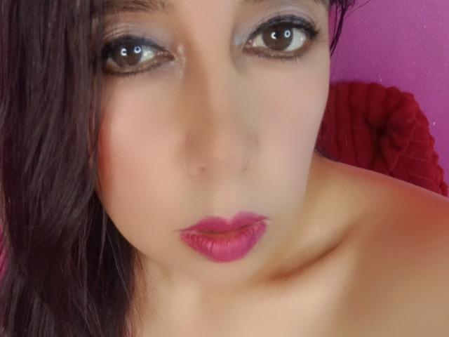SublimeMature has brown eyes, pink lipstick, and a slight frown with dark brown hair partially covering her face. Her bare shoulders are visible. She looks serious.
