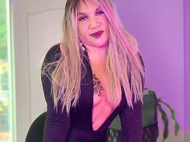 Mia Mancini hat blondes Haar, violette Augen und dunkelroten Lippenstift. Sie trägt ein schwarzes Oberteil mit tiefem V-Ausschnitt, das ihre Brüste und ein Tattoo zeigt. Sie lehnt sich vor und lächelt..