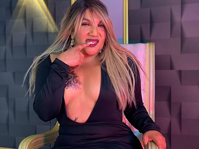 Mia Mancini mit blonden Haaren, tiefschwarzem Kleid, das ihre Brüste zeigt, mit Finger im Mund, Tätowierung auf der linken Brust, sitzend auf einem Stuhl, sexy, lächelnd..