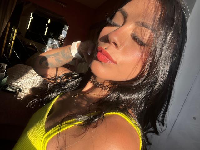 Amberv, com cabelo preto brilhante, blusa amarela, batom vermelho, piercing no nariz, colar, braço tatuado e rosto iluminado pelo sol, olhos fechados, sorrindo, usando um relógio branco, sob a luz do sol..