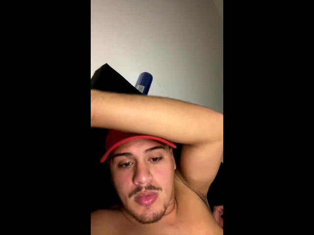 Gabrielzao, shirtless, with red cap, pouty lips, and arm raised, showing a tattoo on his bicep. Background is dark, wall is light. Blue chair in upper background.