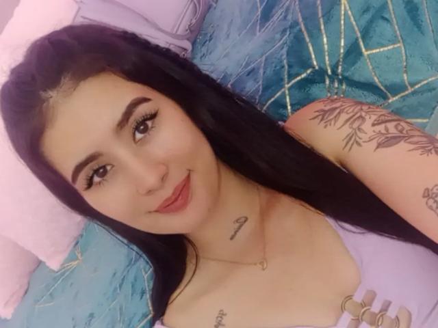 AlisonKweel has black hair, brown eyes, and a small smile. She's wearing a purple top, showing tattoos on her chest and arm, with a visible necklace. Boobs visible.