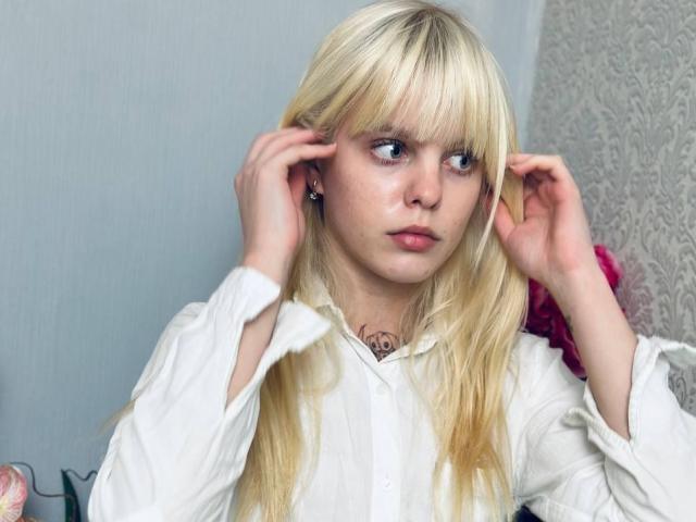 Lialit heeft blond haar, blauwe ogen en draagt een witte blouse. Ze raakt haar oren aan met beide handen en kijkt er iets verward uitzien. Een kleine tatoeage is zichtbaar op haar nek..