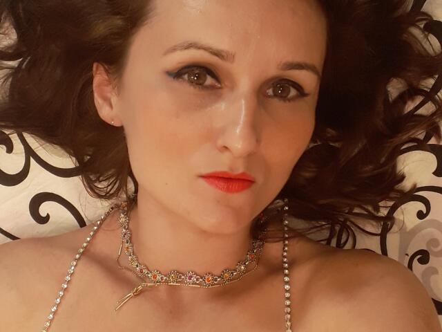 LamourCestMoi has red lipstick, dark makeup, and a shiny silver necklace with a small decorative piece, lying down with wavy brown hair, looking at the camera.