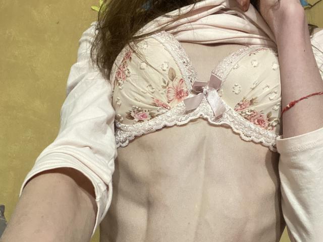 Os seios de Kabblasha estão em um sutiã rosa, adornado com flores e fitas, levantando uma camisa branca para revelar seu torso musculoso, pálido, ligeiramente magro e com o abdômen plano, com uma pulseira vermelha..