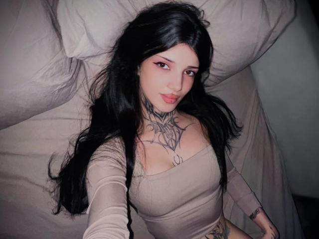 Rianka with long black hair, pink eyeliner, and tattoos, lying on a bed, wearing a beige, off-shoulder top, showing her small tits and a tattoo on her chest.