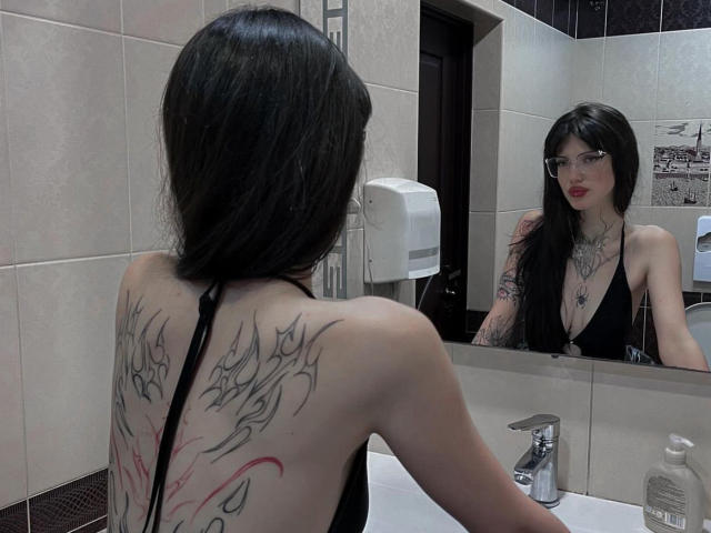 Rianka with long black hair, glasses, and a black halter top, showing a tattoo on her back. She's standing in front of a mirror, looking at herself.