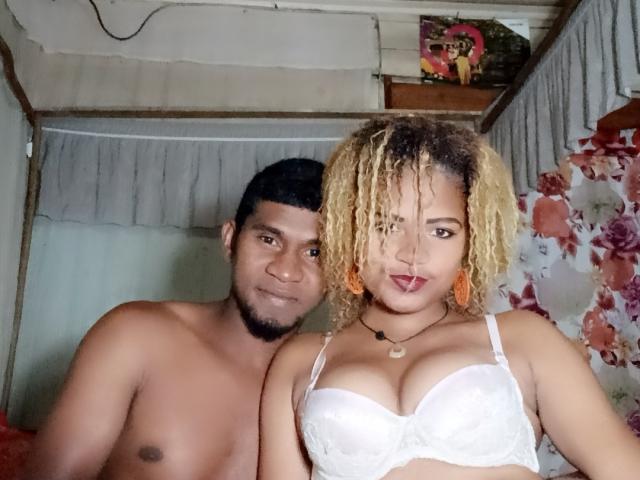 MeriAndMark with dark skin, short black hair, and stubble, shirtless, standing next to MeriAndMark with light brown skin, curly blonde hair, white lace bra, tongue out.