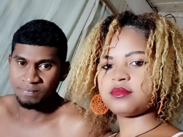 MeriAndMark with curly blonde hair and orange earrings, wearing a white bra, stands next to MeriAndMark with short black hair and a beard, both shirtless. Boobs, tits, pussy, etc.