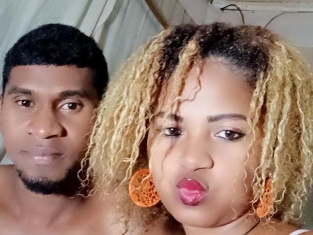 MeriAndMark has curly blonde hair, red lipstick, and orange earrings. MeriAndMark's black-haired, shirtless partner with a goatee stands next to them. Both have dark brown skin.