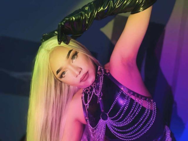 CharlotteCollens with long blonde hair, black latex gloves, and a purple chain-studded top, posing with a raised arm, showing her tits and a seductive look.
