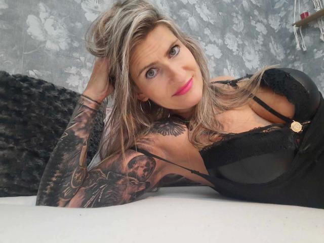 KatyStar-hot with tattoos on her arm and chest, lying on a bed in black lingerie, showing her tits, with pink lipstick and a slight smile.