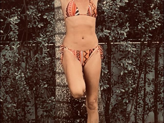 BlackAngeel's slim, tan body with small tits and a cute ass is wearing a red, patterned bikini, standing under a shower with water droplets visible.