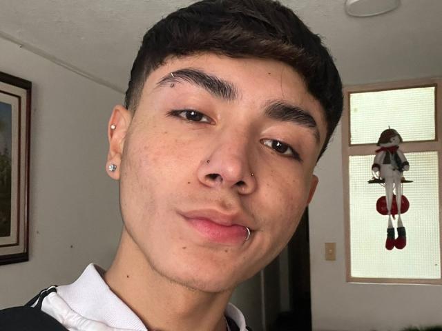 ImJuanda has a nose ring, lip ring, and two ear piercings. Short dark hair, brown eyes, light skin. Small scar above right eyebrow. Doll on window in background.
