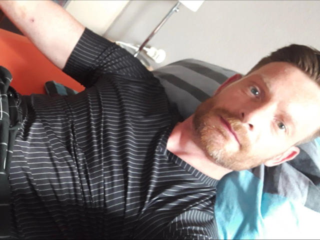 Defender-hot, a red-haired dude with a beard, wearing a black striped shirt, laying on a bed, looking at the camera with a slight smile, and blue pillow.