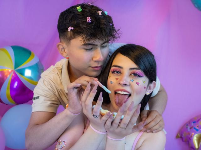 NoahAndRose with curly brown hair and pink star clips, painting colorful makeup on Rose's face, who has pink eyeshadow, white face paint, and her tongue out. NoahAndRose has a beige shirt.