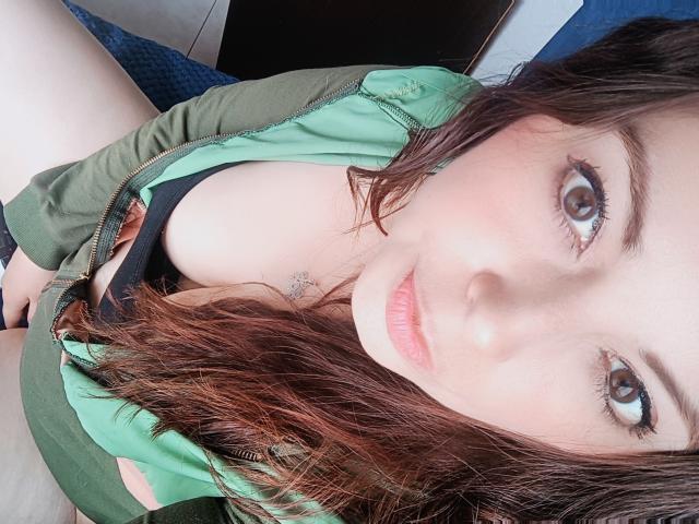 Seher with brown eyes, green zip-up, slightly open, showing black bra and cleavage, wavy brown hair, light skin, small nose, subtle pink lips, sitting with legs apart.
