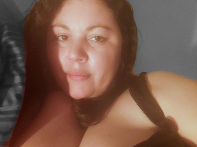 ExoticEva with light brown skin and dark brown hair, wearing a black strap top, has soft brown eyes and a slight smile, showing her bare shoulders and chest.