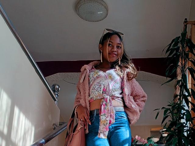 MayhZ with medium brown skin, wearing a pink fluffy jacket, floral tied top, and blue jeans, standing in a sunlit room, holding a pink bag, smiling.
